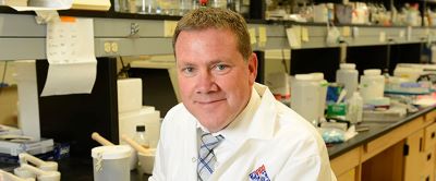 man in lab coat sitting at lab station looking at camera