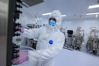 person in PPE including white hood and suit, blue mask, safety glasses and pink gloves works on machine with tubes